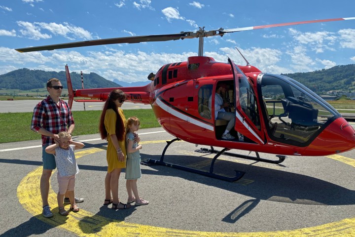 Family standing near a red helicopter on a sunny day with mountains in the background.