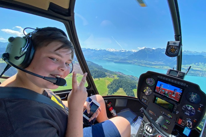 Person in helicopter cockpit with headset, making a peace sign, overlooking a lake and mountains.