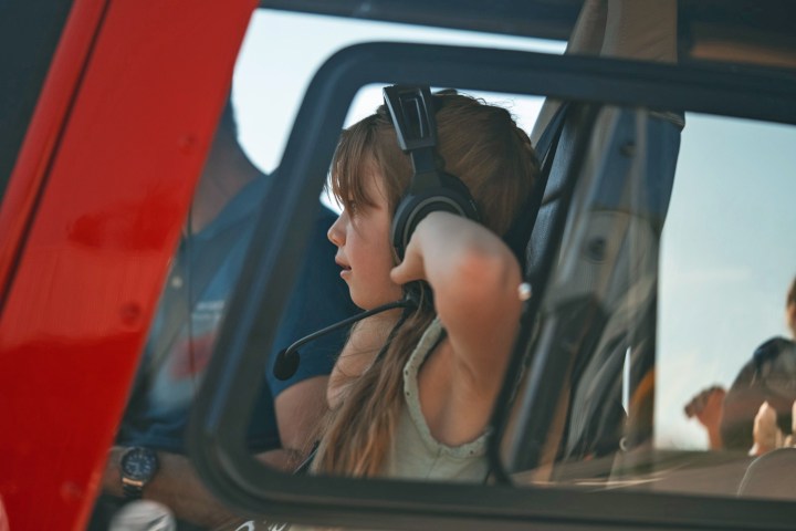 Child with headset sits inside a vehicle or cockpit, looking forward through the window.