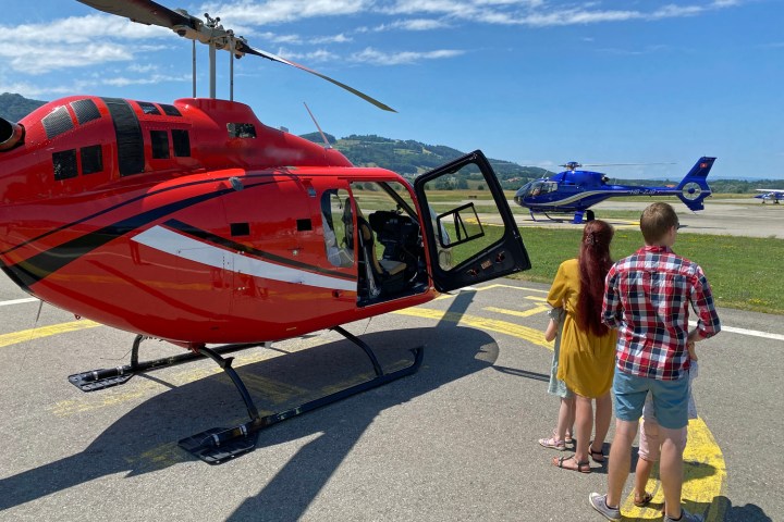 Two people near a red helicopter, with a blue helicopter in the background on a sunny day.