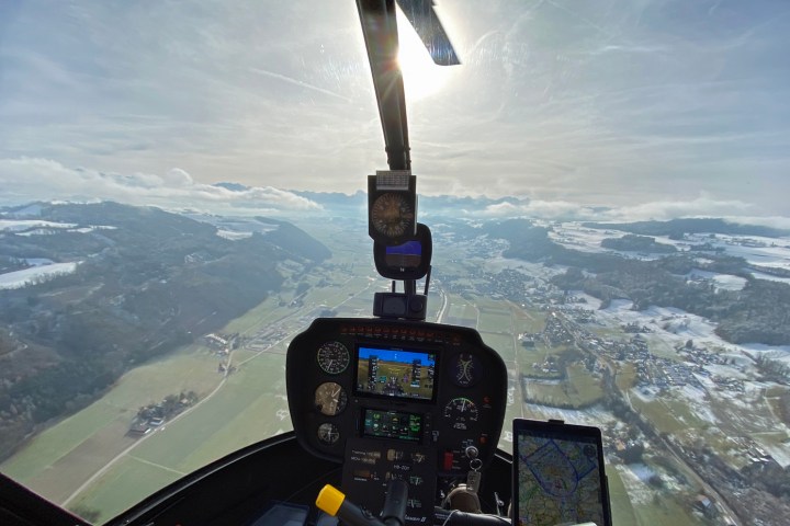 Helicopter cockpit view flying over snowy hills and fields under a sunny sky.