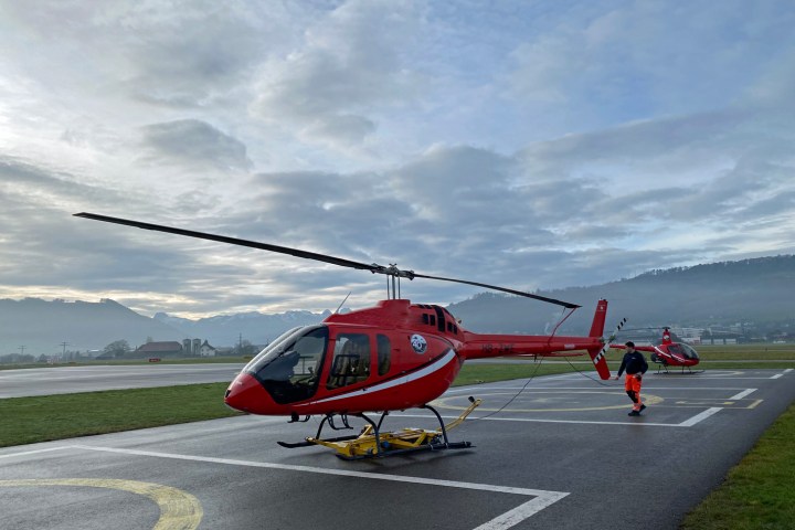 Two red helicopters on an airfield with a person nearby, cloudy sky in the background.