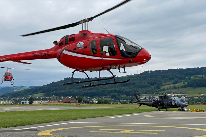 Two red helicopters hover near a helipad with a black helicopter on the ground, against a hilly landscape.