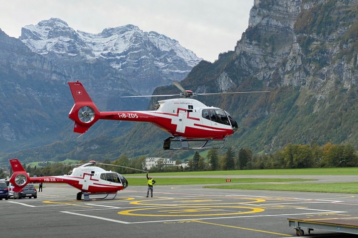 Two red and white helicopters on a mountain helipad, one in the air, with scenic mountain backdrop.
