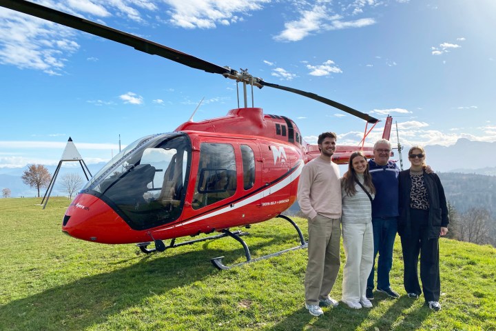Four happy passengers standing next to a red helicopter on a grassy hillside during a scenic stopover at a mountain restaurant in Switzerland, with the Alps in the background.