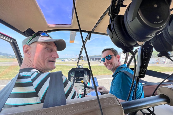 Smiling passenger seated in the co-pilot seat next to a helicopter pilot preparing the cockpit instruments before take-off for a scenic flight over the Swiss Alps.