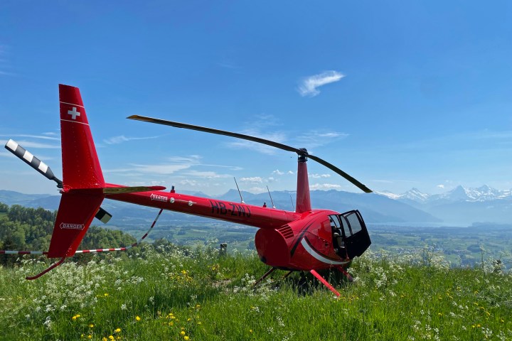 Robinson R44 helicopter parked on a grassy alpine hillside during a scenic stopover in Switzerland, while passengers walk toward a nearby mountain restaurant.