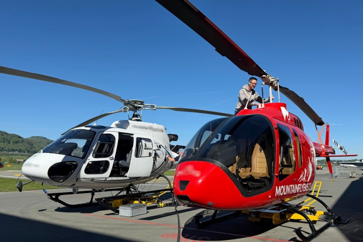 Pilot conducting pre-flight inspection on red helicopter while another helicopter is being refuelled at the station.
