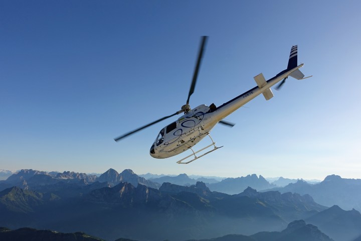 White and blue Airbus H125 helicopter flying high above a mountain valley surrounded by alpine peaks.