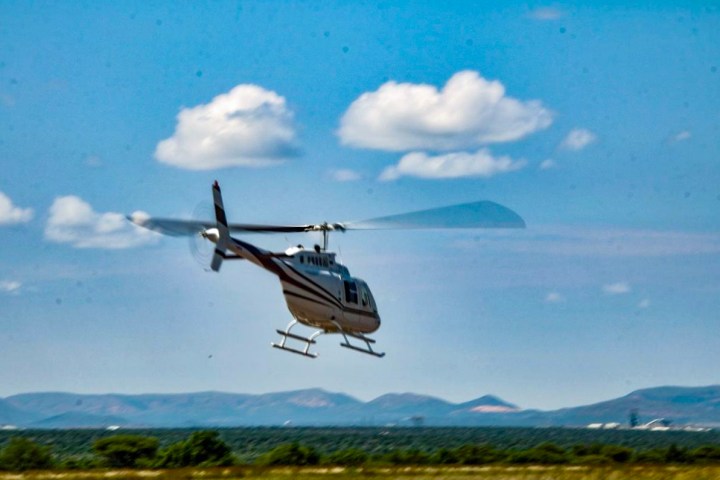 White Bell helicopter flying quickly over green fields under a blue sky with small hills in the background.
