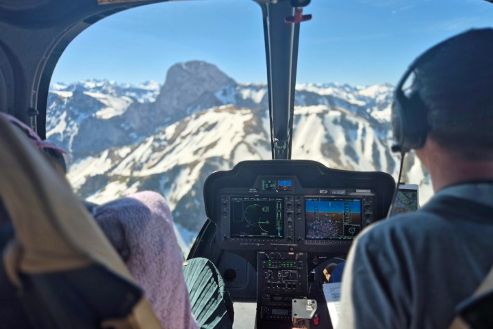 View from the back seat of a helicopter showing a modern digital cockpit, pilot flying over snowy Swiss mountains, and passenger taking photos through the window.