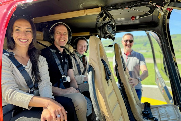 Three happy passengers seated inside a Bell 505 helicopter before a scenic flight over the Swiss Alps while the pilot provides safety instructions at Bern-Belp Airport.