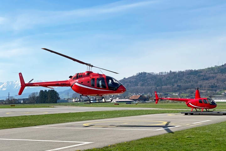 Red Bell 505 helicopter flying above the landing area while a second helicopter waits on the helipad at Bern-Belp Airport during FunFlights scenic helicopter operations.