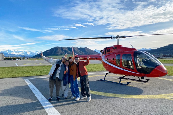 Four excited passengers posing beside a red Bell 505 helicopter at Bern-Belp Airport before their private FunFlights helicopter tour to the Swiss Alps.