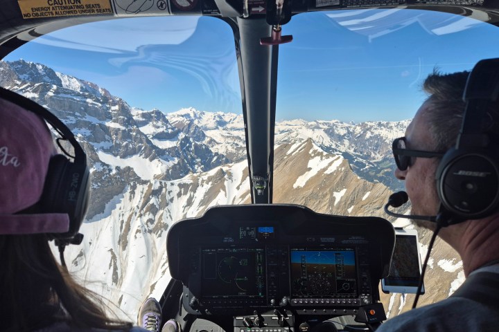 Pilot explaining the Swiss Alps to a passenger from the modern Bell 505 cockpit with snow-covered mountain peaks visible outside.